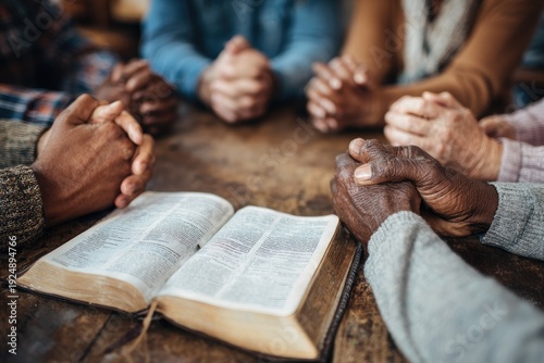 Diverse hands clasped in prayer around a wooden table with an open book