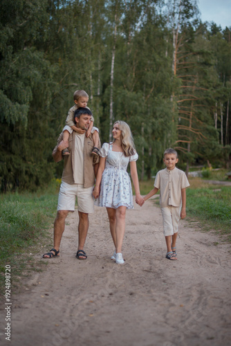 A family walks along a dirt path in a forest. The father is Asian, carrying a young boy on his shoulders. A Caucasian woman and a young boy walk beside them.