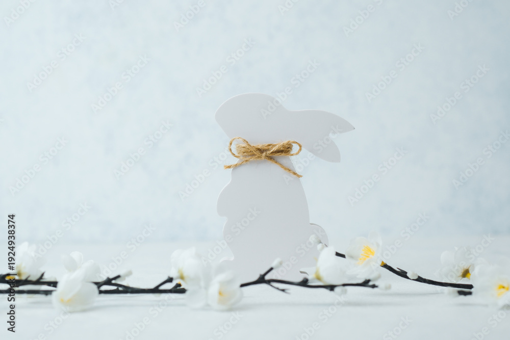 Fototapeta premium Elegant white Easter bunny cutout with twine bow and white flowers on bright background