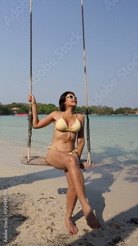 A woman in a yellow bikini and sunglasses poses while sitting with legs crossed on a wooden rope swing over a sandy beach
