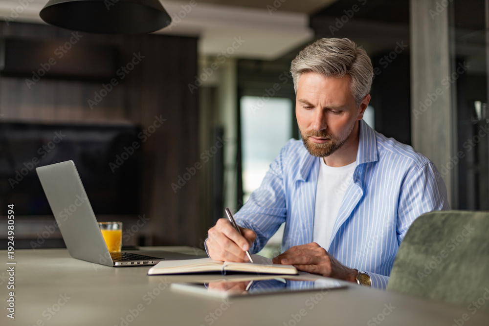 Obraz premium Young businessman sitting at home working on a laptop and writing notes down on a notepad.