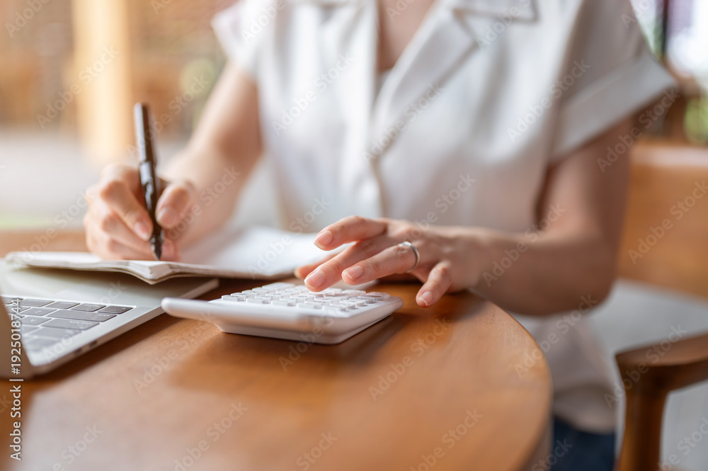 Fototapeta premium Close up of a woman's hand writing in book as using calculator while sitting at wooden table in cafe