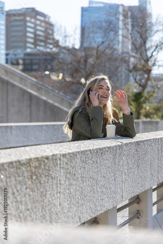 Woman in 20s leaning on railing at terrace, talking on smartphone, olive green jacket, takeaway cup