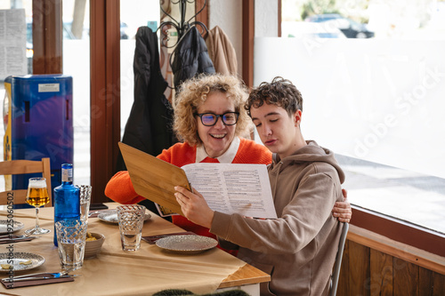 Middle aged mother and teenage son reading restaurant menu together and smiling while choosing food at table