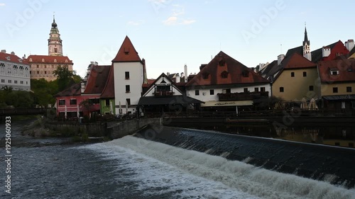 High-angle panoramic view of Cesky Krumlov during sunset. The footage captures the historic castle, the iconic round tower, and the red-roofed medieval houses nestled along the Vltava river.