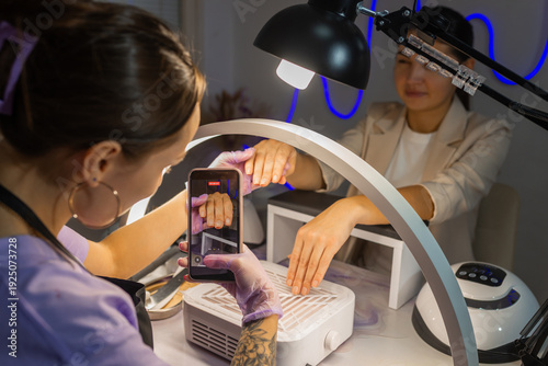Nail technician photographing finished manicure on female hands using smartphone under ring lamp in beauty salon. Professional nail care, content creation, social media and manicure service concept.