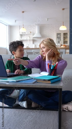 Positive cute African American schoolboy and caring attractive teenage nanny studying lesson online using laptop pc, discussing educational material at home.