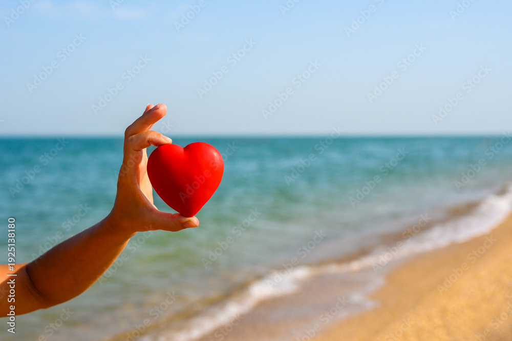 Fototapeta premium A red heart in a hand against the backdrop of the sea. A wonderful seaside vacation.