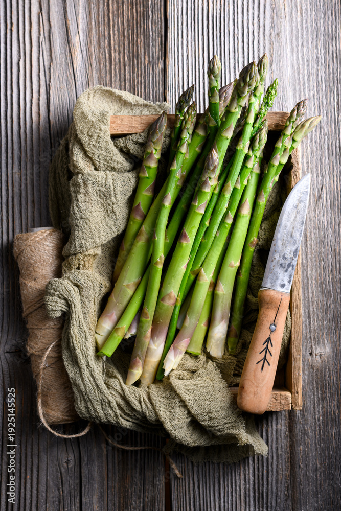 Fototapeta premium Fresh green asparagus bundle arranged with fabric napkin and knife on wooden background. Natural raw vegetables in food photography style. Flat lay, top view