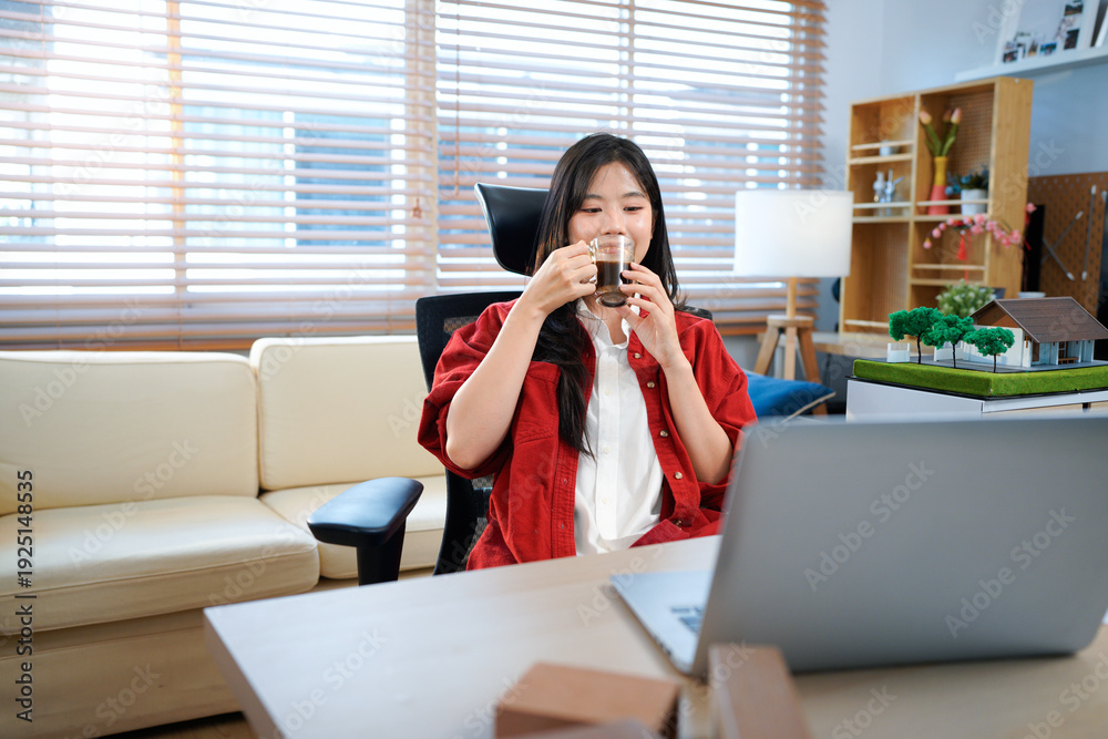 Obraz premium Young architect designer works at home in a cozy atmosphere. Smiling Asian female architect drinking hot coffee at desk with blueprints in a cozy home office, enjoying a cup of coffee