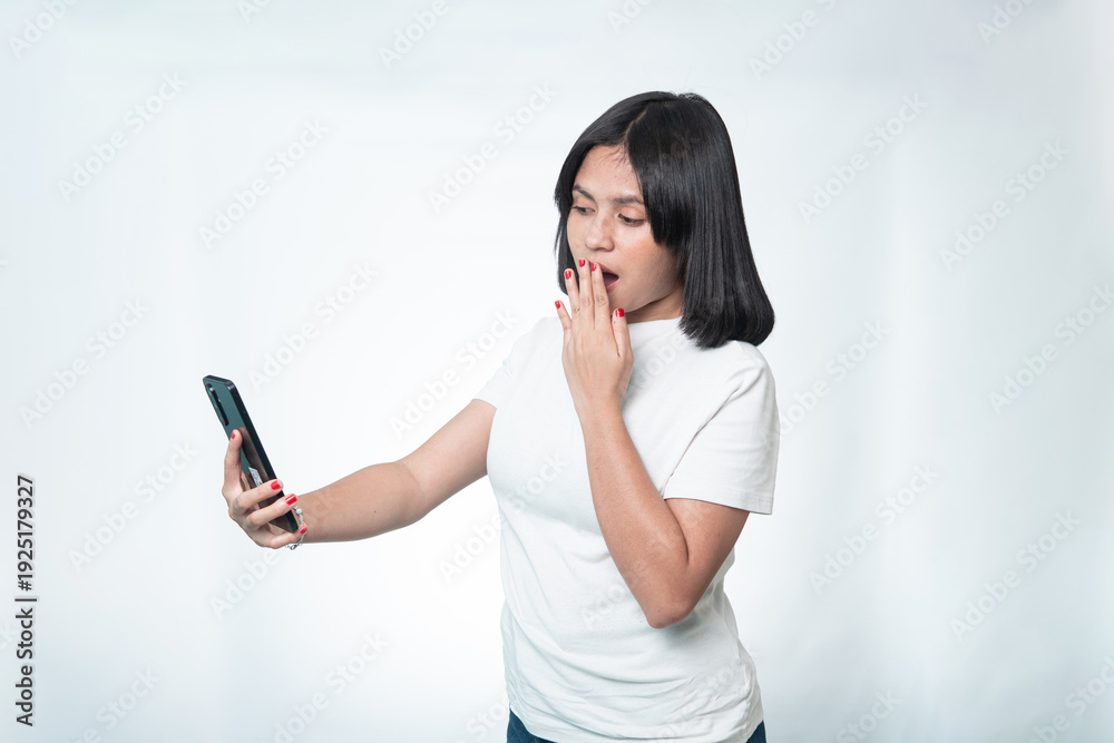 Fototapeta premium Portrait of a shocked Indonesian woman in a white shirt seeing amazing news on her phone. Concept of surprise, disbelief, and viral content. 