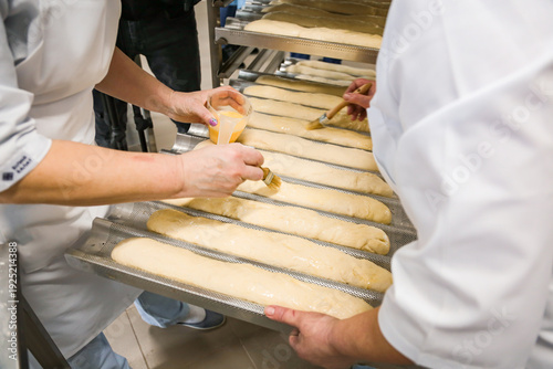 Women brushing baguettes with egg wash in a bakery kitchen before baking. Traditional bread preparation process in professional food production environment.