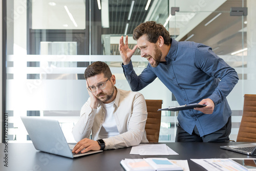 Male boss screaming and shouting at a frustrated male employee working on a laptop, creating a tense atmosphere of stress, pressure, and conflict in the modern office