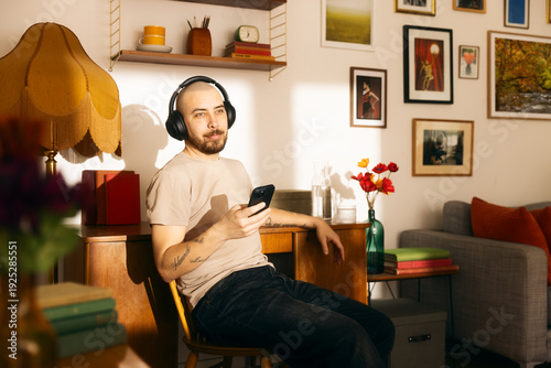 A relaxed man with a shaved head and tattoos sits in a sunlit, vintage-style living room, wearing over-ear headphones and looking at his smartphone while enjoying music or a podcast.