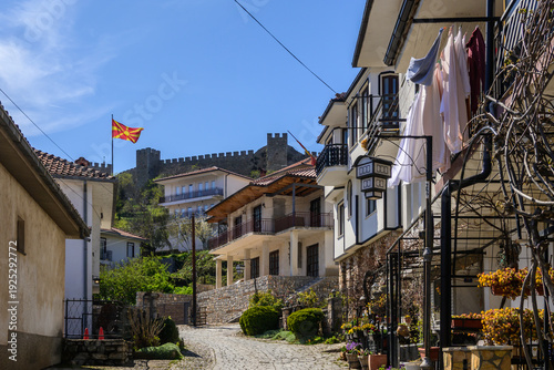 Canvastavla A charming cobblestone street winds through traditional Ohrid houses, leading towards Samuel's Fortress, with a vibrant Macedonian flag fluttering prominently overhead under a sunny sky