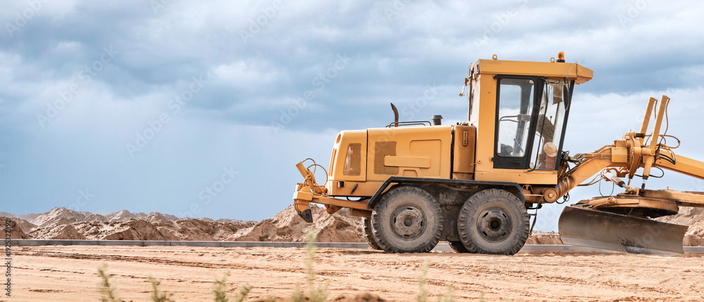 Obraz premium A yellow grader is actively leveling the sandy terrain at a construction site while dramatic clouds loom in the sky during late afternoon