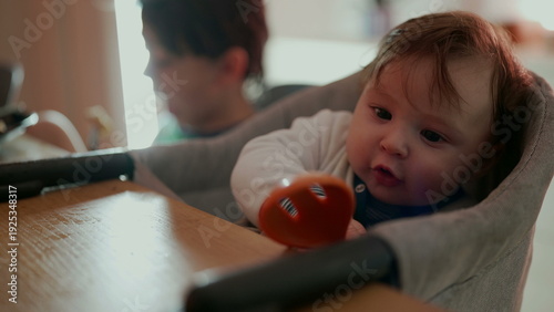 Photography Baby holding orange spoon with wide-eyed expression, joyful sibling in backgroun