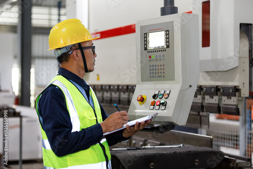 An Asian man, an industrial engineer by profession, wearing a safety helmet, operates machinery and records inspection logs in a factory.