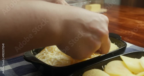 Chef sprinkles grated parmesan cheese over thinly sliced potatoes arranged in baking dishes. Close-up of food preparation, homemade potato casserole, rustic kitchen, natural light, traditional recipe