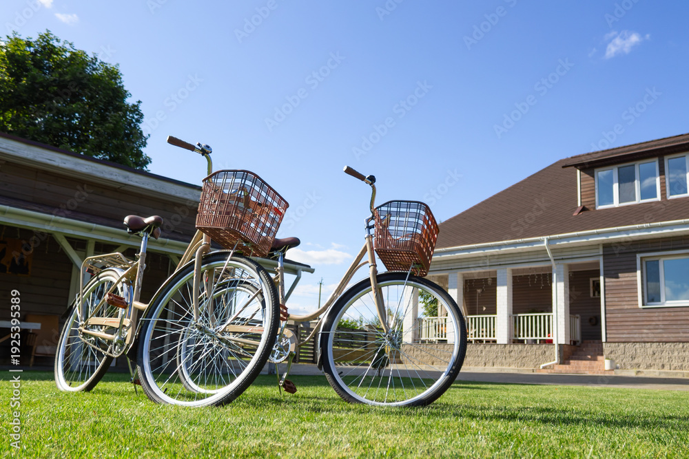 Fototapeta premium A pair of vintage bicycles with wicker baskets parked on a green lawn in front of a modern wooden house on a sunny day. Summer leisure concept with two bicycles near a country house