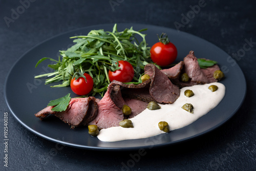 Vitello Tonnato with arugula, cherry tomatoes, capers, and parsley. A popular Italian appetizer made with veal and a tuna-anchovy sauce. Selective focus, black background, close-up.