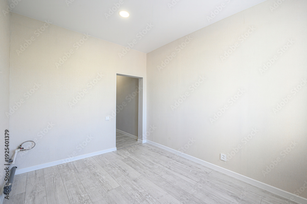 Obraz premium A spacious empty room with light beige walls, grey laminate flooring, a recessed ceiling light, a doorway, electrical outlets, and exposed plumbing pipes