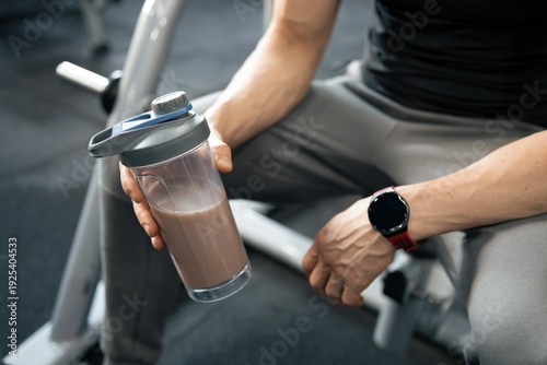 Athletic mature man holding chocolate protein shake after workout in modern fitness club.