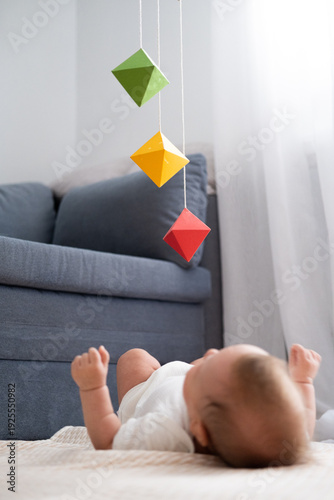 Baby lying on the floor under colorful hanging mobile toys in a modern living room, early development activity, home parenting moment, playful learning.