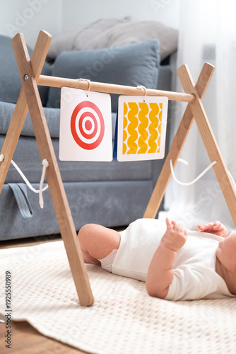 Baby lying under wooden children's play center with hanging colored patterned cards, early learning, home life, playful childhood.