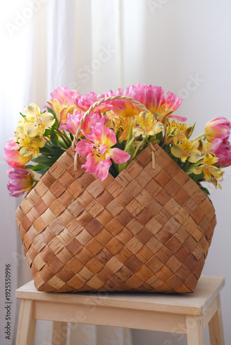 Woven basket filled with colorful flowers on a wooden stool in a bright room, home decor detail, spring interior styling, natural decoration, springtime festivity.