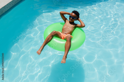 African youth male reclining on green inner tube in blue pool wearing orange trunks and shades