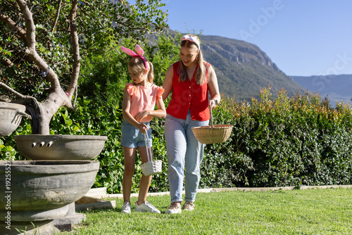 Female adult, child walking on lawn by planter holding baskets wearing red top and bunny ears