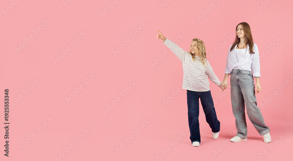 Fototapeta premium Little girl pointing forward on pink studio background and holding mothers hand, expressing curiosity and positive family lifestyle moment, copy space