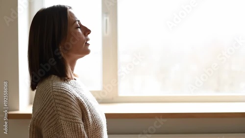 Woman practicing deep breathing exercises by a window. Side profile of female meditating with closed eyes. Mindfulness and mental wellness concept. Copy space for text