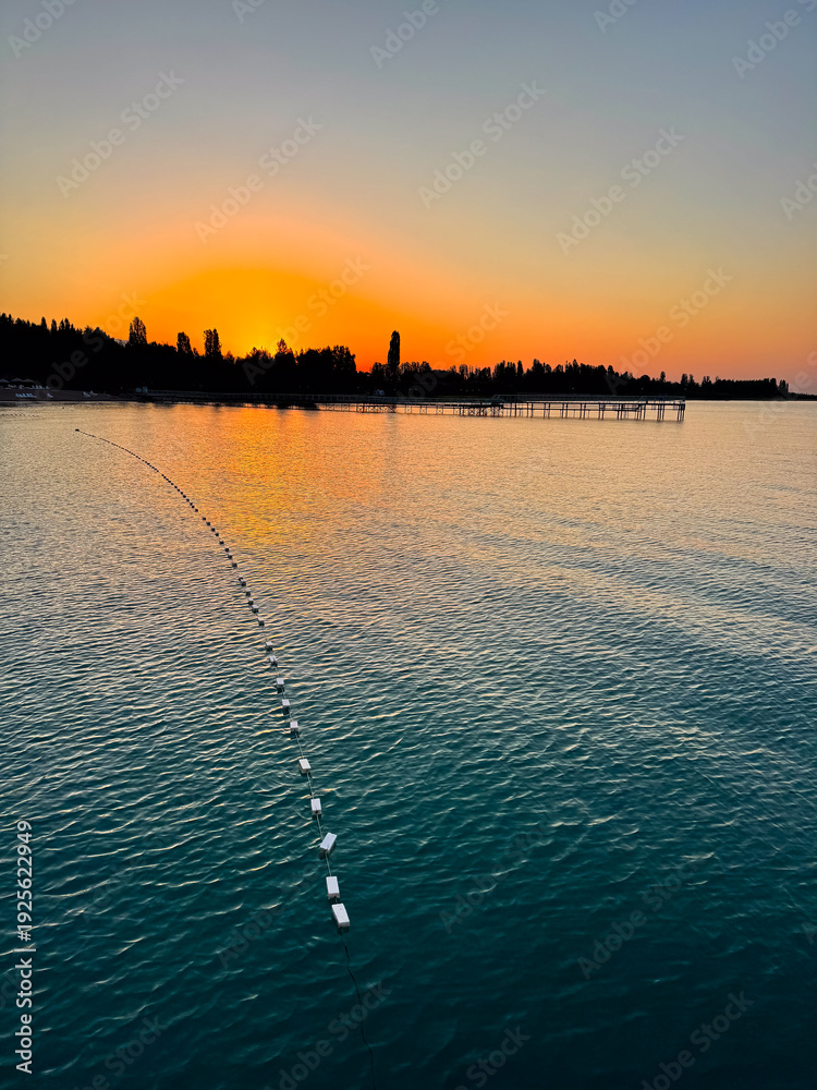 Fototapeta premium Early sunrise at Issyk Kul Lake in Kyrgyzstan. Warm golden sky reflecting in calm water with silhouettes of trees and pier on horizon.