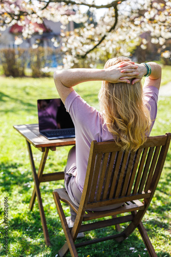 Woman relaxing with hands behind head while working on laptop outdoors in spring garden