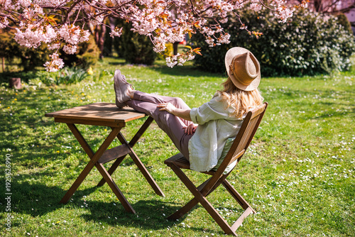 Woman relaxing in spring garden under blooming cherry tree with feet up on table. Enjoyment of sunny day outdoors