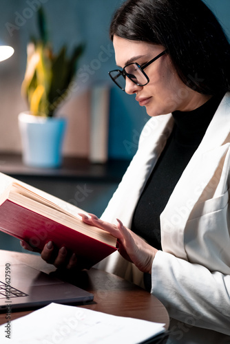 Focused businesswoman reading reference book at office desk, preparing materials for client presentation. Professional preparation, productivity and office workflow concept.
