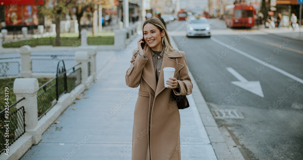 Obraz premium Woman talks on phone while walking on city street with coffee in hand during day