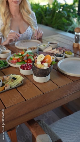 Elegant outdoor brunch spread featuring fresh fruit salad, cheese, and charcuterie with a woman in the background