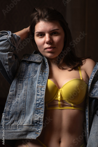 Young woman with long dark hair wearing a yellow bra and denim jacket poses in low light, showcasing her features and the texture of the clothing