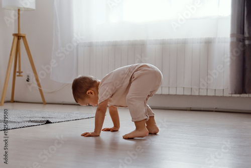 A baby crawls on hands and knees on a wooden floor in a bright room filled with sunlight. Soft light filters through the curtains