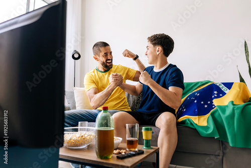 Brazilian father and teenage son watching football match together at home celebrating exciting moments. Family bonding through sports entertainment sharing emotion, support and generational connection