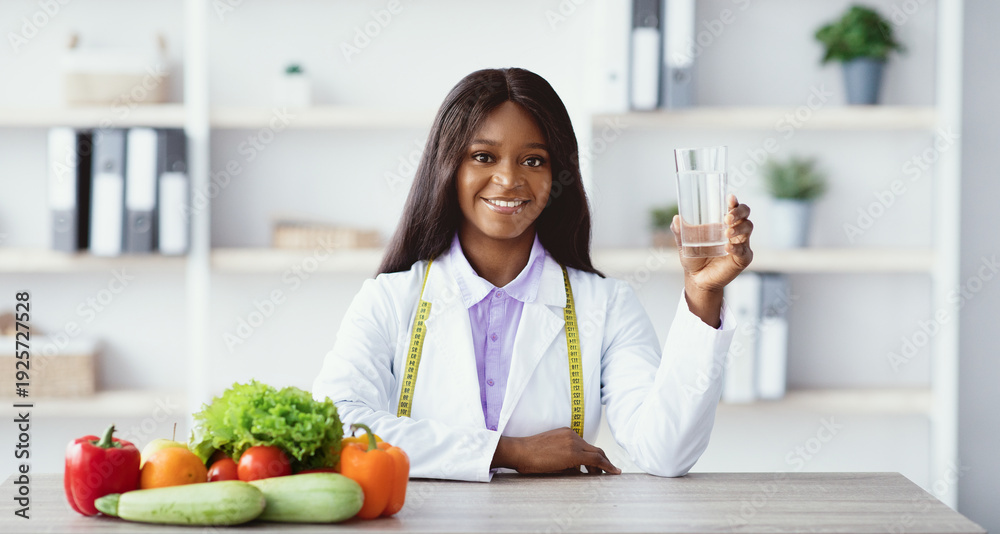 Fototapeta premium A woman in a lab coat holds a glass of water while sitting at a table with various fresh vegetables and fruits. She is promoting a healthy lifestyle in a bright office setting.