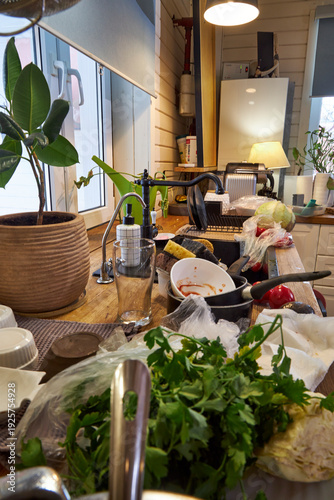 Messy modern kitchen after cooking with dirty dishes, fresh vegetables and cluttered countertop. Open dishwasher and black sink in realistic home interior. Everyday domestic life and household chaos 