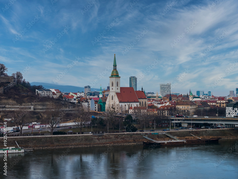 Fototapeta premium St. Martin's Cathedral with green and gold spire rises over Bratislava. Danube River, bridges, embankments, modern high rises, and winter bare trees appear in cool daylight.