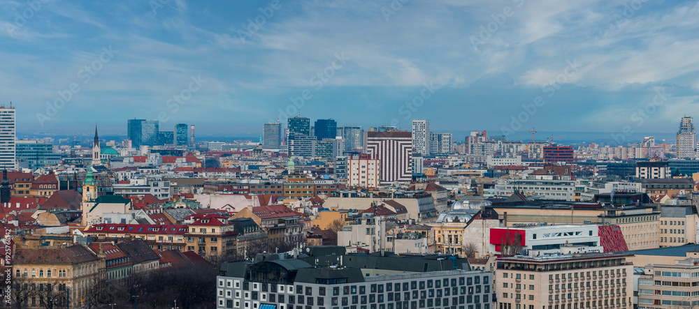 Obraz premium Aerial view of Bratislava, Slovakia, shows historic rooftops, the green capped Old Town Hall tower, church spires, and modern glass high rises under a bright partly cloudy sky.