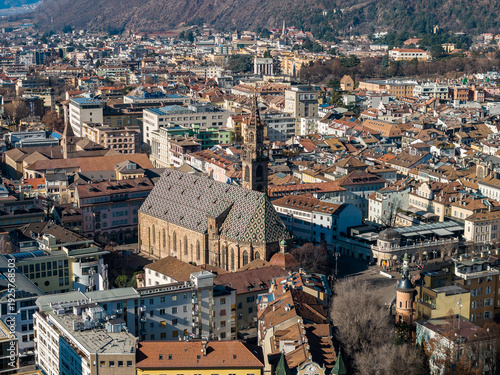 Photography Aerial view of Bolzano, Italy, shows Bolzano Cathedral with patterned roof and spire, pastel and terracotta roofs, wooded hills, and crisp midday winter light