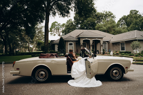 High angle cinematic portrait of a fashionable bride and groom posing inside a vintage convertible with red leather seats. 