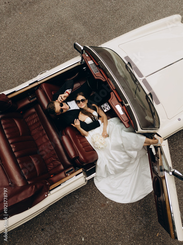 High angle cinematic portrait of a fashionable bride and groom posing inside a vintage convertible with red leather seats. 